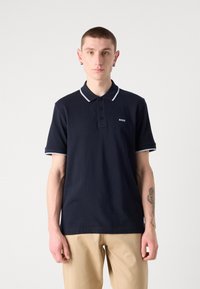 Mornarska polo majica s tridelno zapenjanjem, belim ovratnikom in majhnim belim logotipom na levi strani prsnega dela, v kombinaciji z svetlo bež hlačami.