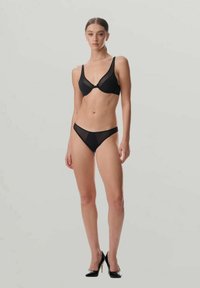 Frau steht mit schwarzem transparenter Büstenhalter und passender Unterwäsche sowie schwarzen High Heels vor grauem Hintergrund.