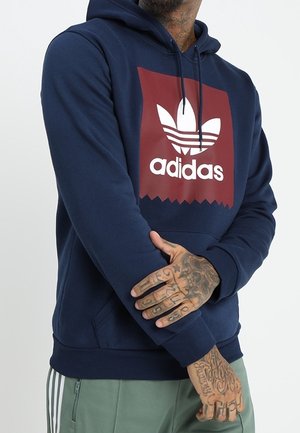 Mężczyzna w granatowej bluzie z kapturem Adidas z czerwonym logo, zielonych spodniach i widocznymi tatuażami na rękach, krzyżując ramiona.