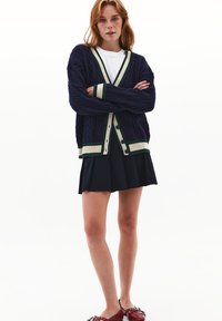 Cárdigan de punto de cable en color azul marino con rayas verdes y crema, de ajuste holgado, con cuello en V y cierre de botones, usado sobre una camiseta blanca y una falda negra.
