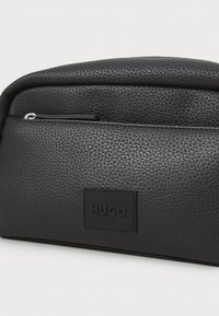Black textured leather pouch with a rounded top, featuring a front zip pocket and a rectangular logo patch embossed with "HUGO."