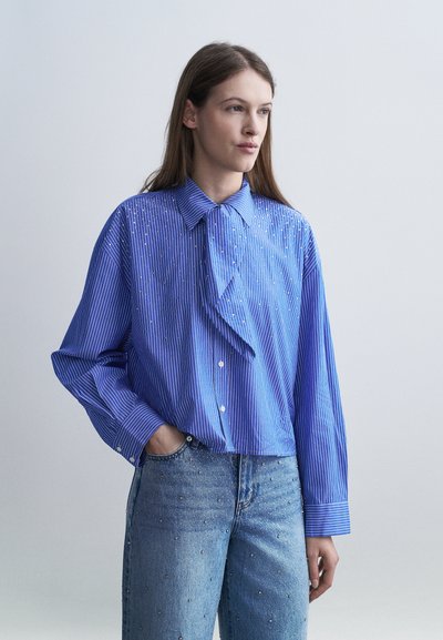 Donna che indossa una blusa a maniche lunghe a righe blu con strass e jeans azzurri decorati con piccoli strass, in piedi con una mano in tasca.