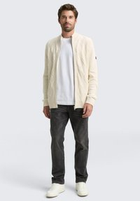 Cremefarbene Rippstrick-Cardigan mit Kabelmuster, mit Reißverschluss, getragen über einem weißen T-Shirt und dunklen Jeans, ergänzt durch weiße Sneakers.