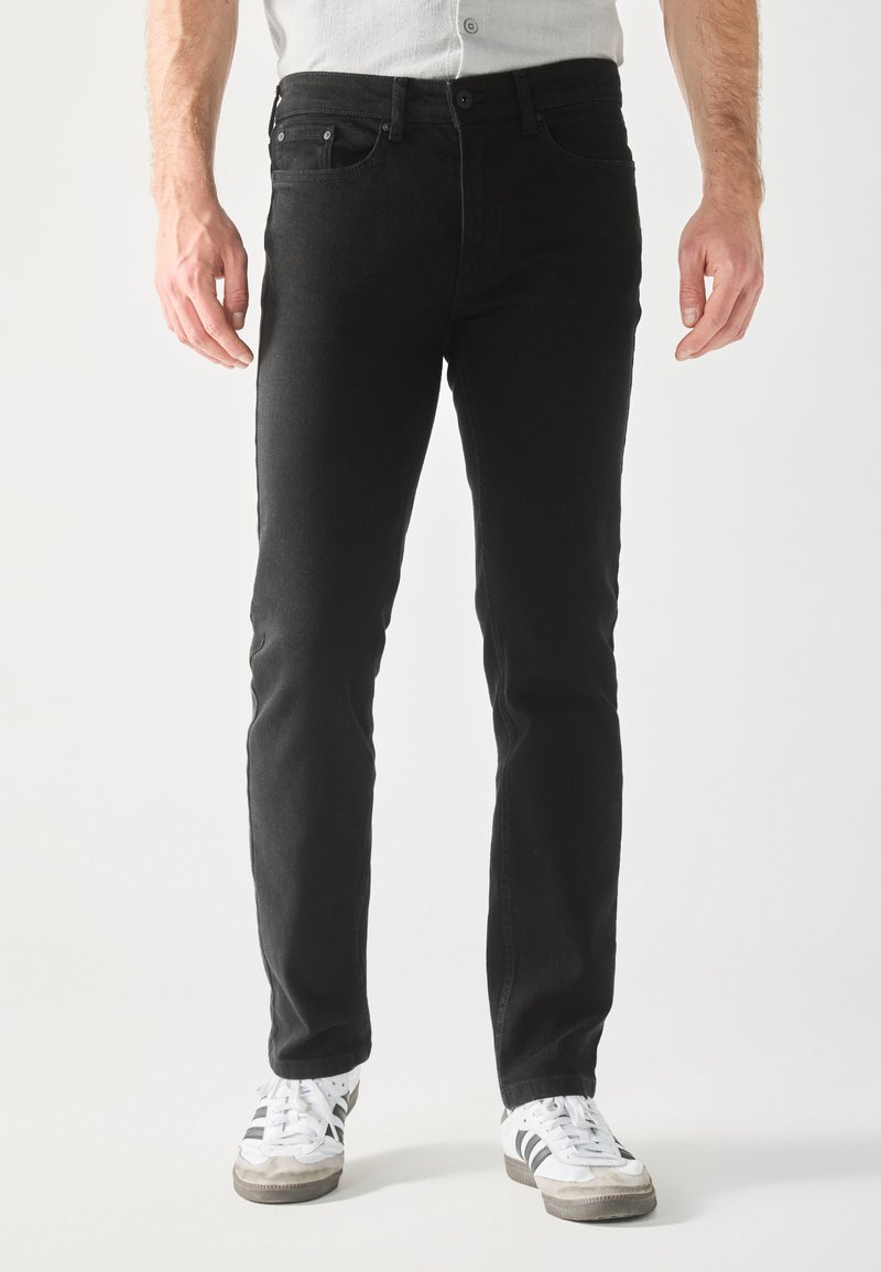 Jeans in denim nero a taglio dritto, cinque tasche e chiusura con bottone. Indossati con sneakers bianche dotate di righe nere.