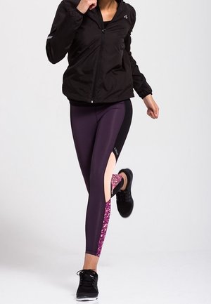 Black zip-up jacket over purple leggings featuring a patterned pink panel and cutouts. Model wears black athletic shoes.