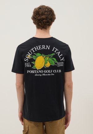 Uomo che indossa una maglietta nera con grande grafica di limone e testo "Southern Italy Positano Golf Club" sulla schiena, in piedi davanti a uno sfondo neutro.