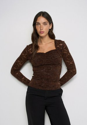 Joven mujer con cabello largo y oscuro lleva una blusa ajustada marrón de encaje con mangas largas y pantalones negros, posando frente a un fondo liso y claro.