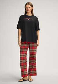 Zwart oversized t-shirt met rood "JOOP!JEANS" logo, gecombineerd met rode geruite broek met zwarte en gouden accenten. Het geheel wordt afgemaakt met slippers.