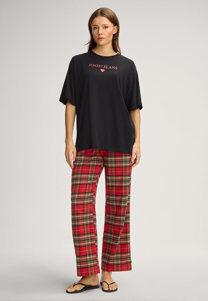 Zwart oversized t-shirt met rood "JOOP!JEANS" logo, gecombineerd met rode geruite broek met zwarte en gouden accenten. Het geheel wordt afgemaakt met slippers.