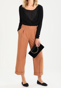 Czarna bluzka z długim rękawem wykończona koronką, brązowe spodnie z wysokim stanem i plisami, czarne płaskie buty oraz czarna kopertówka z okrągłym uchwytem.