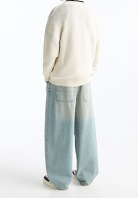 Crème gebreide trui met ribbeltextuur, gecombineerd met lichtblauwe wijde spijkerbroek met een vervaagde gradient. Zwarte afwerking aan de hals van de trui.