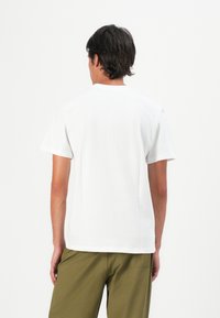 Man wearing plain white short-sleeve t-shirt and olive-green pants, standing facing away against a white background.