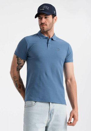 Camiseta polo azul de algodón, con una tapeta de tres botones y un logo bordado. Se lleva con jeans azul claro y una gorra oscura.