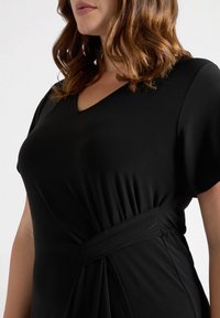 Zwarte jurk met korte mouwen, V-hals en geplooid zijdetail. De stof lijkt glad en rekbaar, wat zorgt voor een getailleerde silhouette.