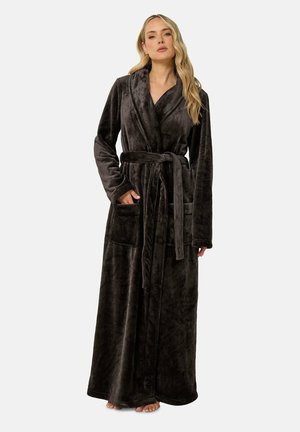 Femme debout portant une longue robe en peluche sombre avec une ceinture nouée à la taille et les mains dans les poches avant, pieds nus sur un fond uni.