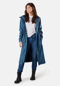 Długi, ciemnoniebieski wodoodporny płaszcz wykonany z błyszczącego materiału. Posiada kaptur, zatrzaski i boczne kieszenie, w zestawieniu z ciemnymi dżinsami i czarnymi butami.