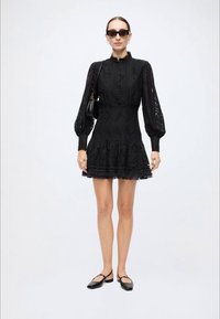 Black lace dress with long, puffed sleeves, a high collar, and ruffled hem. Accessories include black sunglasses and a small handbag.