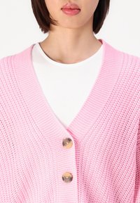 Rosa strikket cardigan med dyp V-hals, med to runde gullknapper, strukturert stoff, og lagd over en hvit topp.