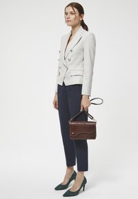 Jasnoszara lniana marynarka podwójnie zapięta, granatowe szyte na miarę spodnie, głęboko zielone szpiczaste szpilki oraz ciemnobrązowa torebka o teksturze.