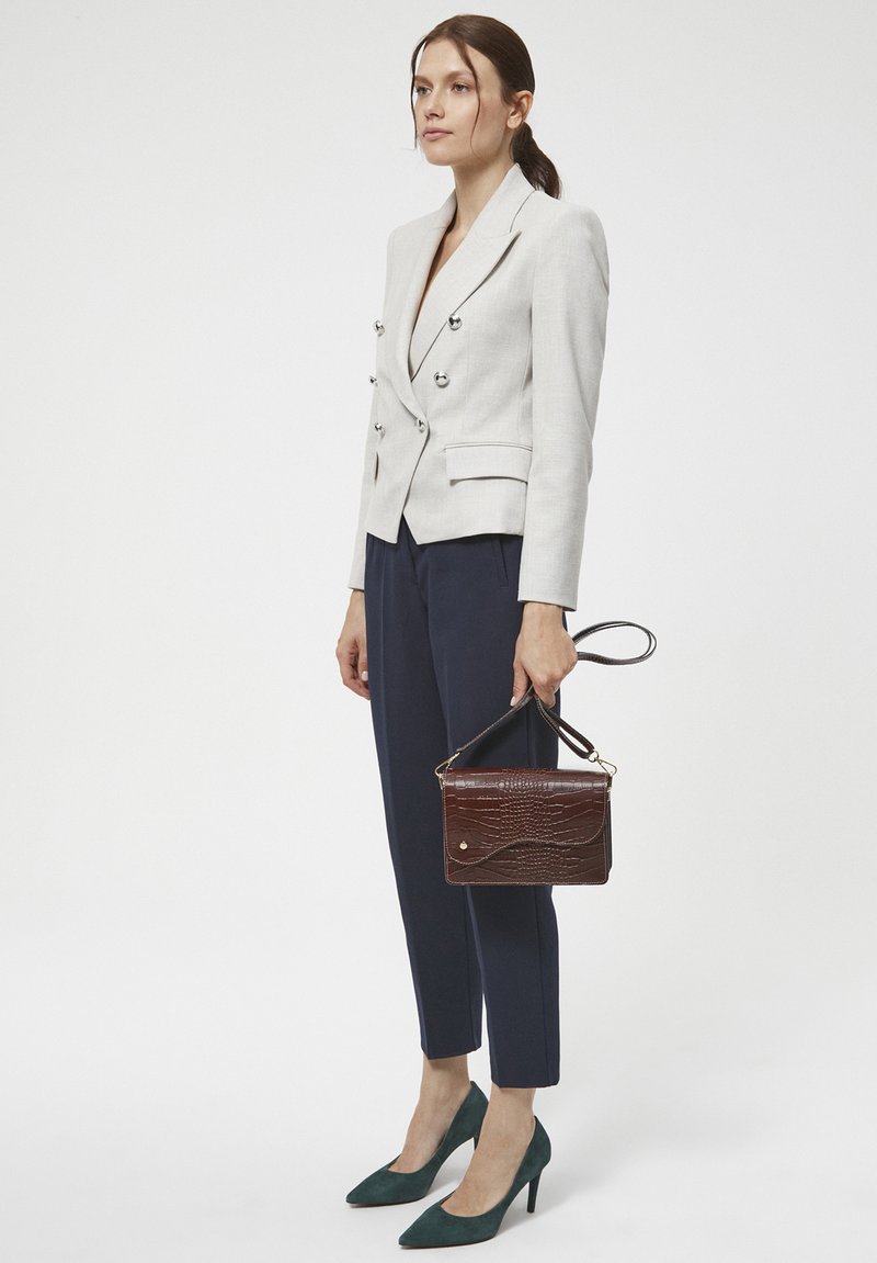 Jasnoszara lniana marynarka podwójnie zapięta, granatowe szyte na miarę spodnie, głęboko zielone szpiczaste szpilki oraz ciemnobrązowa torebka o teksturze.