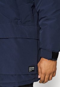 Mörkblå puffjacka med texturerad tyg, med en patchetikett längst ner till vänster och kardborreband vid ärmsluten.