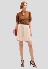 Braunes Strickoberteil mit kurzen strukturierten Ärmeln, kombiniert mit cremefarbenen, hochgeschnittenen Shorts, braunem Gürtel und Riemchenheeledschuhen, während sie eine rote Handtasche hält.