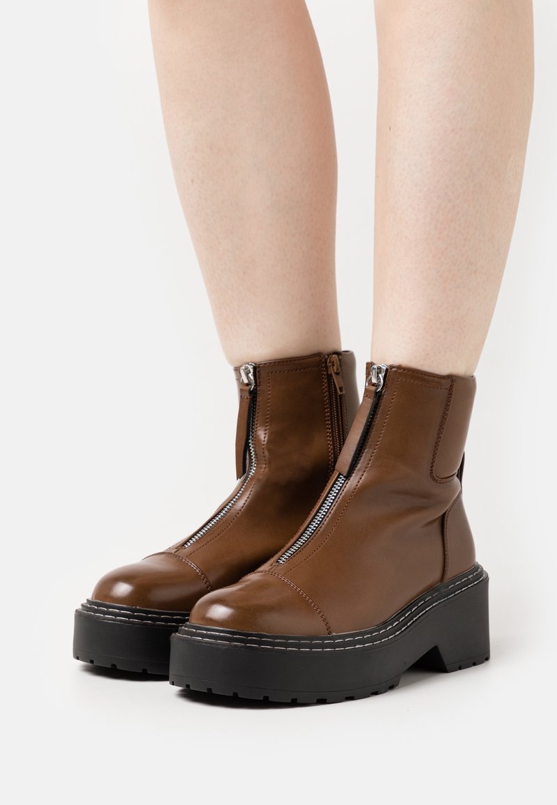 Bottes cheville en cuir marron avec fermetures éclair latérales, semelles compensées épaisses noires et détails de surpiqûres blanches contrastantes.