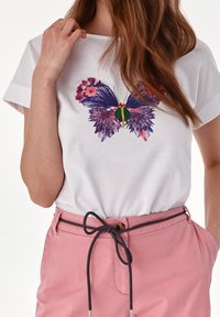 Witte T-shirt met een vlinderprint in paars en florale details. Gecombineerd met roze broeken en een zwarte trekkoordriem.