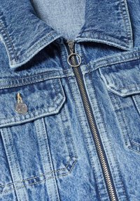 Sininen farkkutakki, jossa on etuvetoketju pyöreällä vetimellä, kaksi rintataskua läpillä ja näkyvissä olevat ompeluyksityiskohdat.