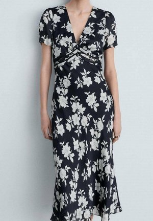 Femme portant une robe noire à motif floral blanc, manches courtes et décolleté en V profond.