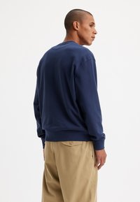Felpa blu navy con girocollo, dotata di polsini e orlo a costine, realizzata in un tessuto morbido. Indossata con pantaloni beige, vista da dietro.