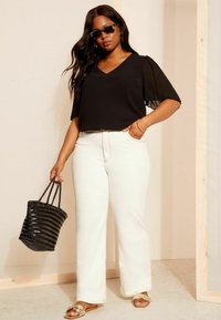 Haut noir avec des manches courtes plissées, jean taille haute blanc, sandales beiges et un sac fourre-tout tissé noir. Fond uni.