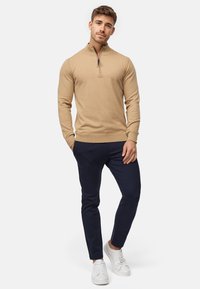 Pull en beige à col zippé avec des poignets côtelés, associé à un pantalon tapered bleu marine et des baskets blanches. Design simple et ajusté avec peu de détails.