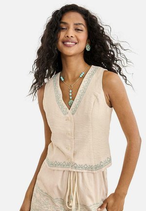 Mujer con cabello rizado lleva una blusa beige sin mangas con botones y pantalones a juego con bordado verde claro y joyas de cuentas turquesas.