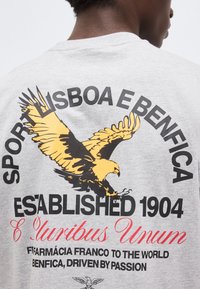 Άτομο που φορά γκρι πουκάμισο με μεγάλο κίτρινο και μαύρο αετό και κείμενο στο πίσω μέρος που γράφει "Sport Lisboa e Benfica, Established 1904, E Pluribus Unum".