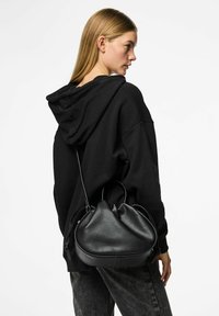 Borsa a mano in pelle nera con forma rotonda e cima arricciata, dotata di manici corti e texture liscia, indossata sopra un hoodie nero.