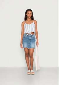 Top cropped listrado azul-claro e branco com laço na frente, combinado com uma saia mini de ganga e sandálias bejes de calçar. Bolsa branca na mão.