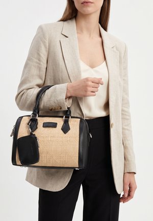 Vrouw in beige blazer en witte blouse die een beige en zwarte gestructureerde handtas vasthoudt met een klein zwart tasje eraan.