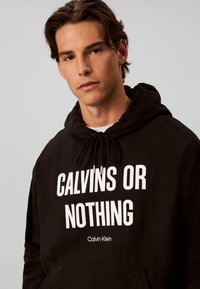 Jeune homme aux cheveux châtain moyen porte un sweat à capuche noir avec un texte blanc indiquant "CALVINS OR NOTHING" et "Calvin Klein" à l'avant.