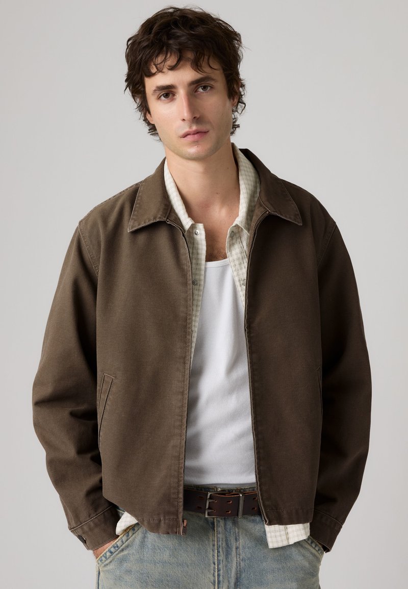 Joven de pie contra un fondo liso que lleva una chaqueta marrón, camisa blanca a cuadros, camiseta blanca y vaqueros azul claro con las manos en los bolsillos.