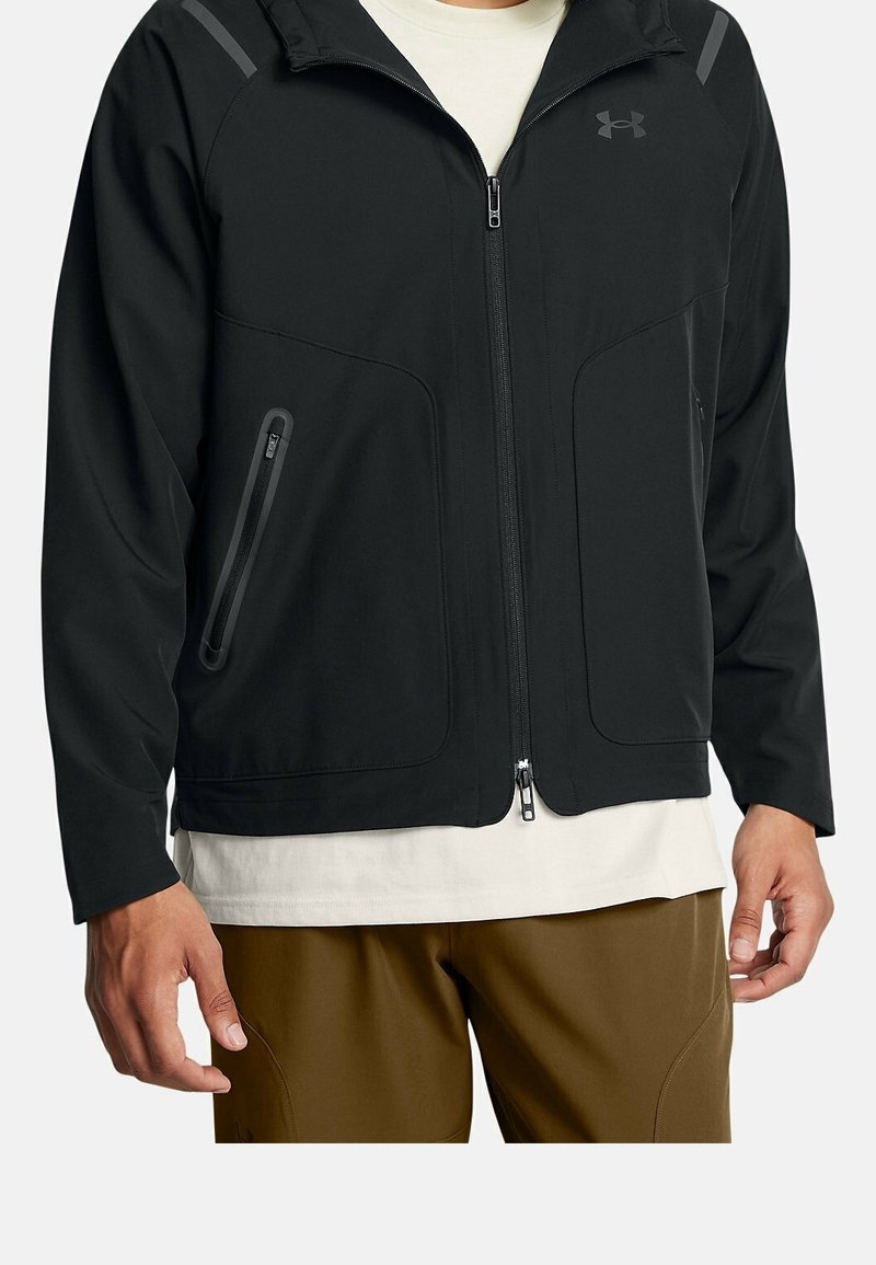 Homme portant une veste noire à fermeture éclair avec un col montant par-dessus une chemise blanche et un pantalon marron, les mains détendues le long du corps.