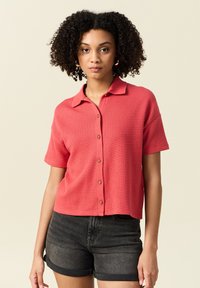 Kortärmad korallskjorta med ett strukturerat stickat mönster, krage och knappstängning, ihop med mörka denimshorts.