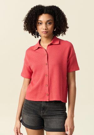 Korte mouwen koraalkleurig shirt met een gestructureerd breipatroon, kraag en knoopsluiting aan de voorkant, gecombineerd met donkere spijkerbroekshorts.