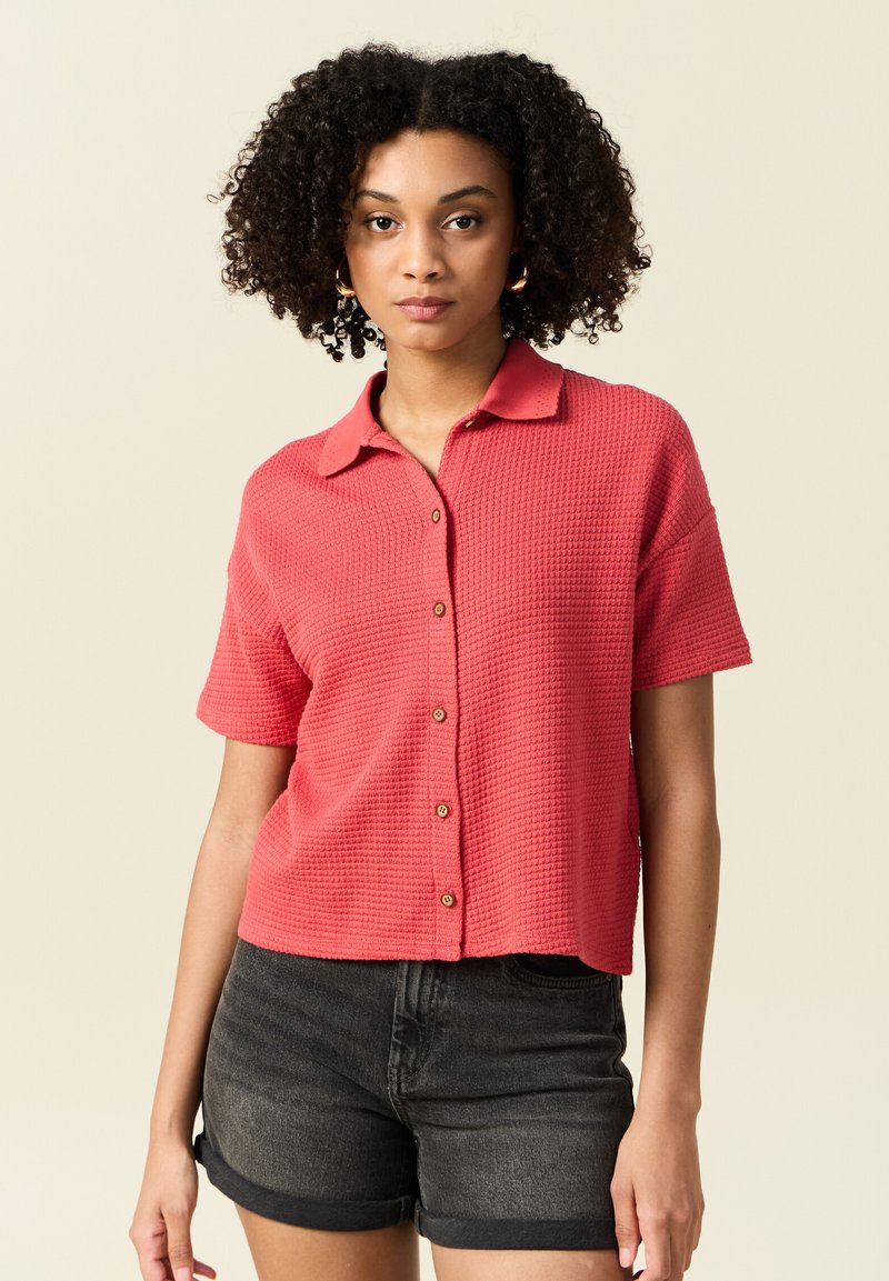 Kortärmad korallskjorta med ett strukturerat stickat mönster, krage och knappstängning, ihop med mörka denimshorts.