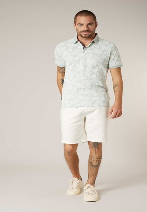 Homme tatoué portant un polo à motifs vert clair, un short blanc et des baskets beiges, debout devant un fond beige uni.