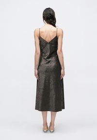 Sieviete ar gariem tumšiem matiem, valkājot bezroku, kroku melnu midi kleitu un bēšas krāsas kurpītes ar zemu papēdi, redzama no aizmugures uz baltas virsmas fona.