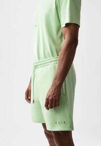 Conjunto deportivo de color verde claro que incluye una camiseta de cuello redondo y mangas cortas, y pantalones cortos con cordón, con detalle de marca bordado. Textura suave.
