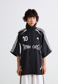 Černá sportovní bunda s vysokým límcem, krátkými rukávy, bílými pruhy, předním zipem a nápisem "TEAM GEIST" v bílé barvě s číslem "10."