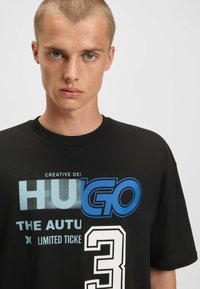Camiseta de algodón negra con un gran texto gráfico azul y blanco, que incluye "HUGO" y "3", con cuello redondo y mangas cortas.