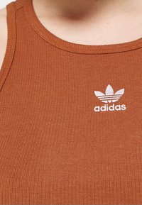 Brązowy, prążkowany top na ramiączkach z okrągłym dekoltem i białym, wyszywanym logo Adidas. Materiał jest gładki z lekkim połyskiem.